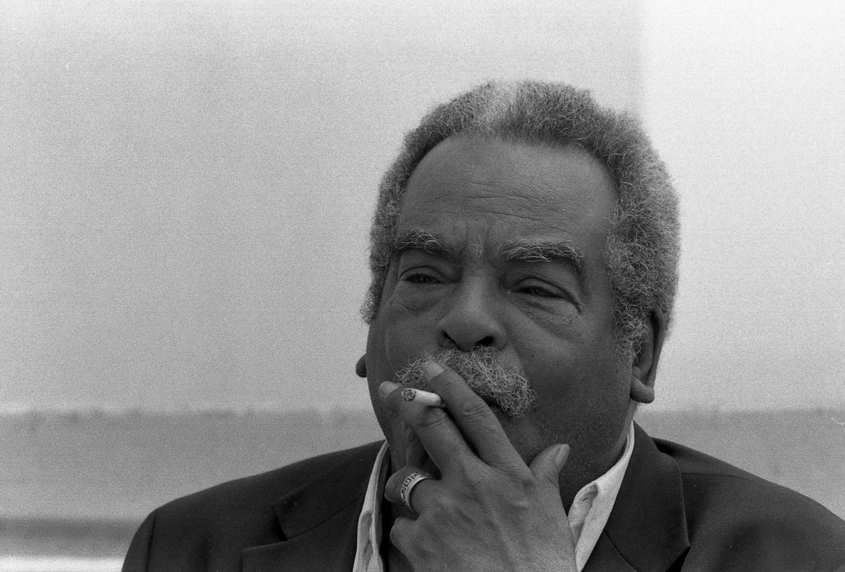 A headshot of Edouard Glissant, who wears a dark suit and smokes a cigarette.