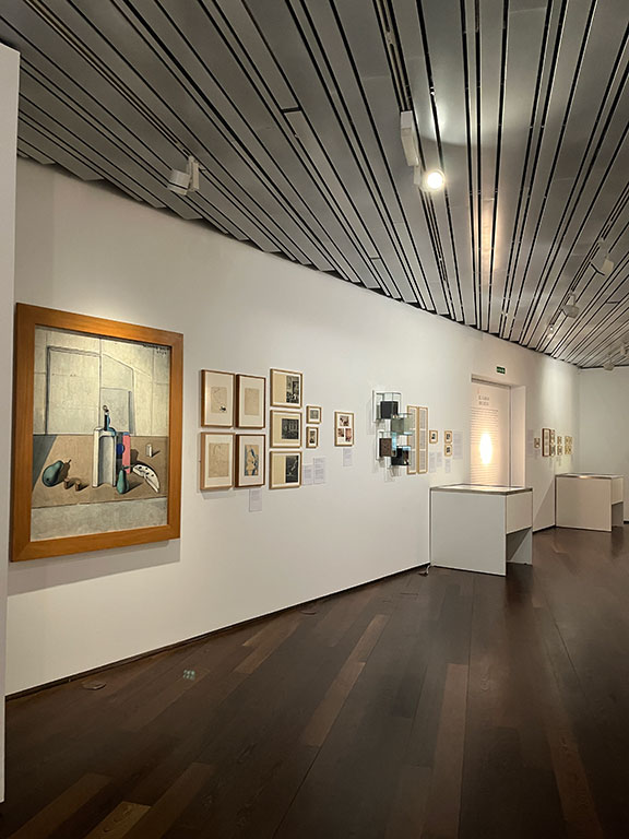 Gallery hallway featuring framed paintings, drawings, and photographs displayed on white walls with track lighting overhead.